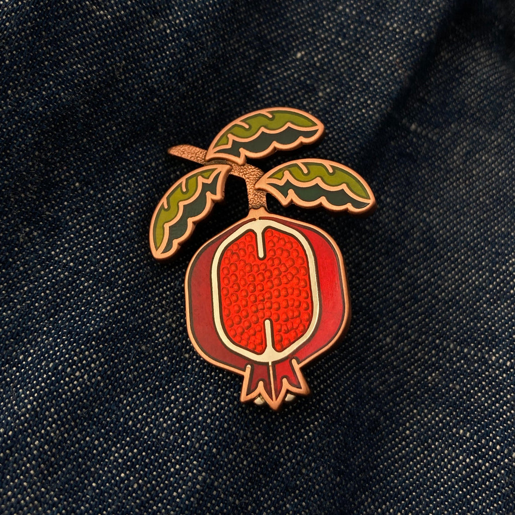 Pomegranate pin seconds in silver and antique copper on a jean jacket with green enamel leaf branch and red sliced fruit.